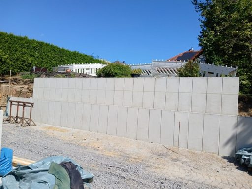 L- Steine Mauer aus Betonblöcken, umgeben von Steinboden und Bepflanzung im Hintergrund.